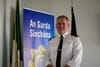 A grey-haired man in a white shirt with epaulettes on the shoulders, and a slim neck tie.